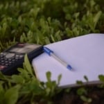 Ein leerer Schreibblock, ein Kugelschreiber und ein Taschenrechner liegen auf einer grünen, kräuterbewachsenen Wiese.