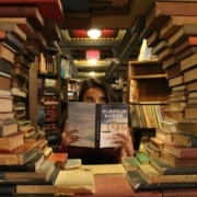Im Vordergrund sind Bücher so gestapelt, dass sie ein kreisförmiges Fenster bilden. Durch dieses Fenster sieht man eine junge Frau, die in einer Bibliothek sitzt. Sie hält sich ein aufgeklapptes Buch vors Gesicht.