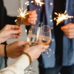 Eine Gruppe von Menschen steht zusammen. Sie halten brennende Wunderkerzen und stoßen mit Champagner auf das neue Jahr an.