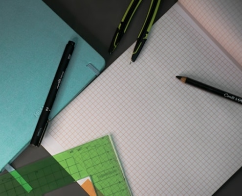 Symbolbild grünes Messen: Ein karierter Block mit mehreren Stiften, einem Zirkel und einem grünen Messdreieck.