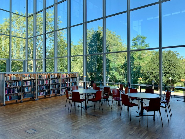 Blick in eine Unibibliothek. Links im Hintergrund Bücherregale, rechts drei runde Tische, umgeben von Stühlen. Der Raum hat eine sehr hohe Decke und verglaste Wände, durch die man einen Garten mit grünen Bäumen und darüber den blauen Himmel sieht.